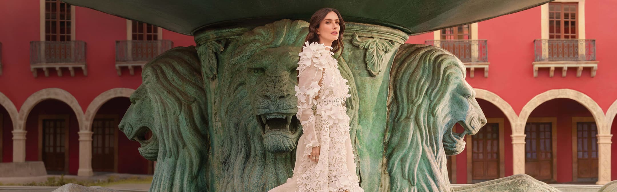  Imagen de una mujer con vestido que parece de novia, con encaje por todos lados y formar de flores, de fondo una estatua que parece fuente color verde con cabezas de leon talladas  al rededor y mas al fondo un edificio color rojo antiguo, UN PALACIO DE LA TALLA DE LEÓN GRAN APERTURA OCTUBRE 2024