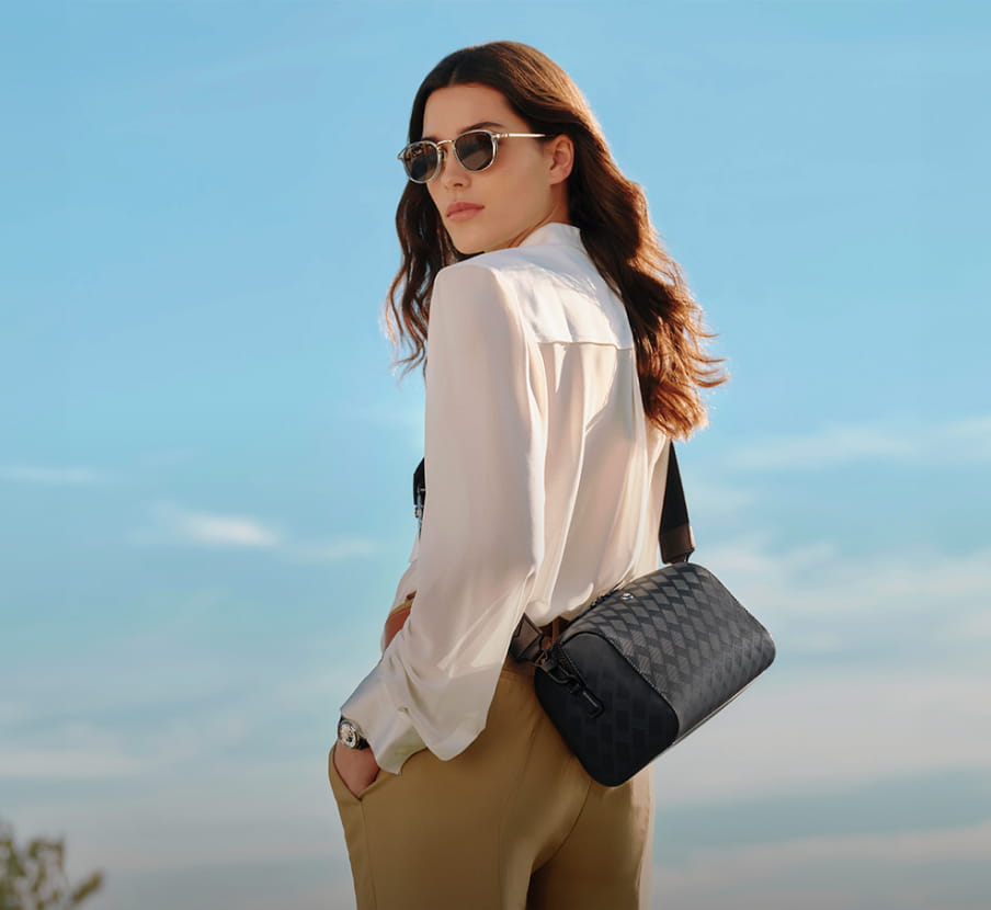 Imagen de mujer con blusa blanca, pantalon cafe, gafas de sol y bolso de piel negro, MONTBLANC